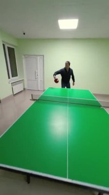Enerjik erkek oyuncu, yeşil masa tenisi masasında dinamik masa tenisi hareketleri ve çeşitli oyun teknikleri sergiliyor. Nane duvarlı iç mekanda sırayla çekiliyor. Dikey Video