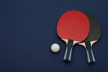 Profesyonel pinpon raketlerinin minimalist kompozisyonu. Kırmızı ve siyah kauçuk yüzeyli, rekabetçi beyaz top, derin donanma arka planında düzenlenmiş.