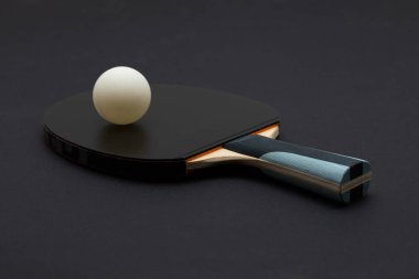 Siyah turnuva sınıfı pinpon raketinin yer aldığı dramatik bir stüdyo kompozisyonu. Parlak ahşap saplı ve beyaz top rekabeti. Koyu renk arka plana karşı zarif bir kontrast oluşturuyor.