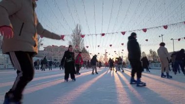 Kalp şeklinde çelenklerle süslenmiş canlı bir açık hava buz pateni pisti. İnsanlar altın günbatımında buzun üzerinde uzun gölgelerle kayarken neşeli ışıklar.