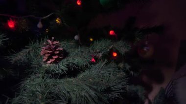 Doğal bir çam kozalağı yeşil bir Noel ağacının dalına uzanır. Etrafı renkli parlak ışıklar ve şenlikli çelenklerle çevrilidir. Sıcak bir tatil atmosferi yaratır.