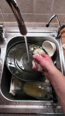 Mutfak lavabosunda akan suyun altında şeffaf bir bardak yıkayan ellerin yakın çekimi. Paslanmaz çelik leğen ve krom musluk temel temizlik rutinini yaparken görünür. Dikey Video