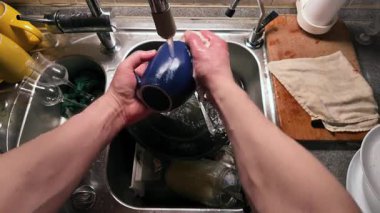 Mutfak lavabosunda akan suyla siyah bir seramik bardak yıkayan ellerin yakın çekimi. Paslanmaz çelik lavabo çeşitli yemekler içerirken ahşap kesme tahtası ve kumaş yakında dinleniyor ve günlük temizlik rutinini gösteriyor.