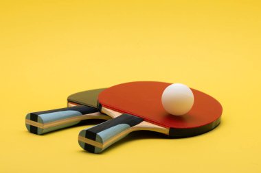Tahta saplı siyah ve kırmızı kauçuk yüzeyli iki profesyonel pin pon raketinin minimalist genel çekimi, canlı sarı yüzey üzerinde rekabetçi sınıf beyaz top eşliğinde
