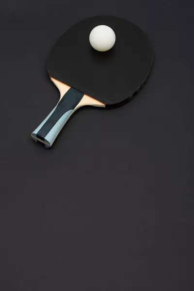 Turnuva derecesinde siyah bir pinpon raketinin minimalist yukarıdan çekimi. Mavi aksanlı ergonomik ahşap kulplu ve koyu arkaplana karşı düzenlenmiş beyaz top.