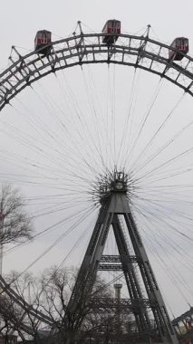 Prater Parkı 'ndaki Viyana Dönme Dolabı, bulutlu bir gökyüzünün altında çıplak ağaçlarla çevrili kırmızı gondolları ve çelik çerçevesiyle dimdik ayakta duruyor. Viyana 'nın tarihi bir simgesi