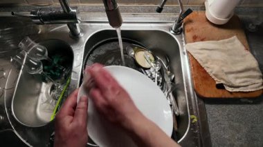 Mutfak lavabosunda bulaşıkları akan suyla yıkayan ellerin yakın çekimi. Paslanmaz çelik lavabo çeşitli cam eşyalar içerirken ahşap kesme tahtası ve kumaş yakında dinleniyor.