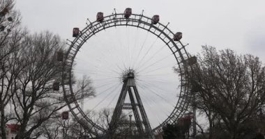 Prater Parkı 'ndaki Viyana Dönme Dolabı, bulutlu bir gökyüzünün altında çıplak ağaçlarla çevrili kırmızı gondolları ve çelik çerçevesiyle dimdik ayakta duruyor. Viyana 'nın tarihi bir simgesi