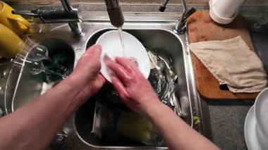 Mutfak lavabosunda akan suyla beyaz bir tabak yıkayan ellerin yakın çekimi. Paslanmaz çelik lavabo çeşitli yemekler içerirken ahşap kesme tahtası ve kumaş yakında dinleniyor ve günlük temizlik rutinini gösteriyor.