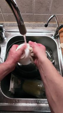 Mutfak lavabosunda bulaşıkları akan suyla yıkayan ellerin yakın çekimi. Mutfak malzemeleriyle dolu paslanmaz çelik lavabo günlük temizlik rutinlerini ve ev işlerini gösteriyor. Dikey Video