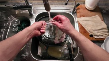 Mutfak lavabosunda akan suyla bardakları yıkayan ellerin yakın çekimi. Paslanmaz çelik lavabo günlük temizlik sırasında çeşitli cam parçalarıyla dolu.