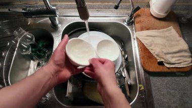 Mutfak lavabosunda akan suyla beyaz bir seramik kabı yıkayan ellerin yakın çekimi. Paslanmaz çelik lavabo çeşitli cam eşyalar içerirken ahşap kesme tahtası ve kumaş yakında dinleniyor ve günlük ev işlerini gösteriyor.