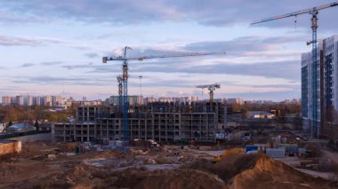 Şehir inşaat sahasında konut iskeletine karşı siluetli vinçler, gün batımında çarpıcı bir şekilde aydınlanan bulutlar ve altın akşam ışığı altında yıkanan toprak parçaları yer alıyor.