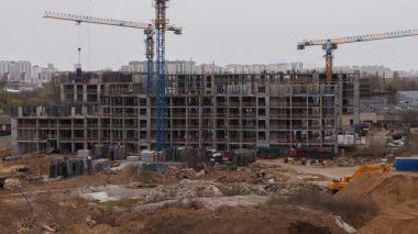 Apartman inşaatı, mavi vinçlerin kaldırdığı devasa beton iskeleti gösteriyor. Ağır makineler inşaat malzemelerini kentsel kalkınma sahasında taşırken, işçiler katlar arasında hareket ediyorlar.