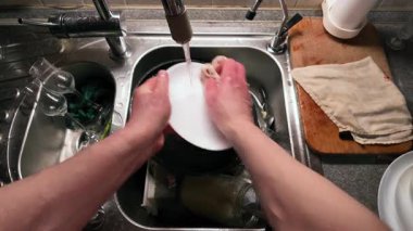 Mutfak lavabosunda bulaşıkları akan suyla yıkayan ellerin yakın çekimi. Paslanmaz çelik lavabo çeşitli yemek takımları içerirken ahşap kesme tahtası ve kumaş yakında dinleniyor ve günlük ev işlerini gösteriyor.