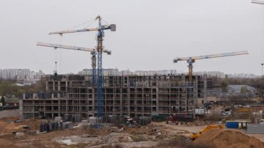 Apartman inşaatı, mavi vinçlerin kaldırdığı devasa beton iskeleti gösteriyor. Ağır makineler inşaat malzemelerini kentsel kalkınma sahasında taşırken, işçiler katlar arasında hareket ediyorlar.
