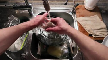 Mutfak lavabosunda akan suyla bardağı yıkayan ellerin yakın çekimi. Paslanmaz çelik lavabo, ahşap kesim tahtası ve kumaş evde temizlik yapıldığını gösteriyor.