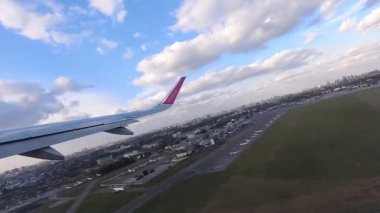 Yolcu penceresinden çekilen belirgin mor kanatlı uçak kanadı şehir manzarası ve havaalanı pistleri üzerinde beyaz bulutlu güzel mavi gökyüzüne karşı.