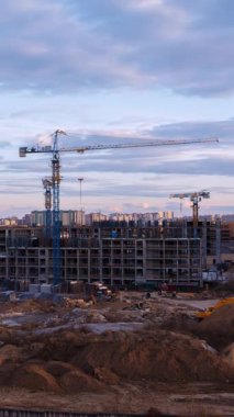 Şehir inşaat sahasında konut iskeletine karşı siluetli vinçler, gün batımında çarpıcı bir şekilde aydınlanan bulutlar ve altın akşam ışığı altında yıkanan toprak parçaları yer alıyor.