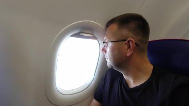 Gözlüklü, odaklanmış erkek yolcunun profil görüntüsü gündüz uçuşu sırasında parlak uçak penceresinden dikkatlice bakıyor, mavi uçak kabininde oturuyor, koltuklarında belirgin kırmızı süslemeler var.