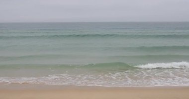 Pembe gömlekli ve açık gri pantolonlu düşünceli bir adam kumlu sahilde duruyor bulutlu gökyüzü altında hafif turkuaz okyanus dalgalarına yaklaşıyor ve huzur dolu bir yalnızlık anı yaratıyor. 