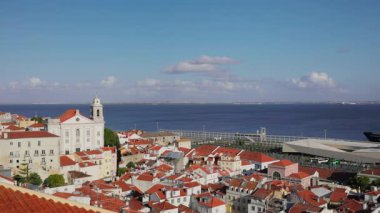 Lizbon 'un tarihi Alfama bölgesinin nefes kesici manzarası. İkonik terasotta çatıları olan geleneksel Portekiz mimarisi ve Tagus Nehri' nin geniş mavi sularına bakan beyaz kilise kulesi berrak yaz gökyüzü altında.
