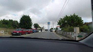 Sesmarias, Portekiz - 13 Ekim 2019: Sesmarias, Lagoa, Algarve, Portekiz 'de köy evleri, park edilmiş arabalar ve bulutlu gökyüzü ile su kulesinin yakınındaki hareket halindeki bir arabadan sokak manzarası