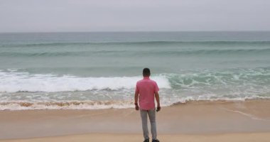 Pembe gömlekli ve açık gri pantolonlu düşünceli bir adam kumlu sahilde durmuş turkuaz okyanus dalgalarının bulutlu gökyüzünün altında sürekli dalgalanmasını seyrediyor.