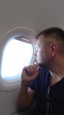 Düşünceli erkek yolcunun dikey portresi gündüz uçuşu sırasında oval uçak penceresinden bakarken, uçak kabininden manzarayı izlerken çenesini dinlendirirken.