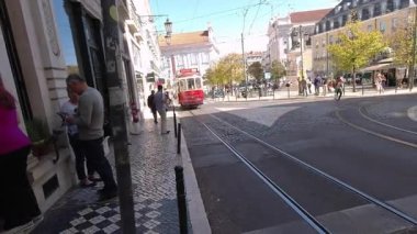 Lizbon, Portekiz - 14 Ekim 2019: Lizbon 'un tarihi bölgesindeki kaldırım taşı caddesinde geleneksel Portekiz binaları ve sarı sonbahar ağaçlarıyla birlikte beyaz bir teslimat minibüsü ile birlikte yol alan ikonik kırmızı klasik tramvay.