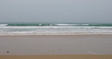 Portekiz 'in Peniche plajında sisli bir günde okyanus dalgalarında sörf yapan maceraperest sörfçüler gri bulutlu gökyüzü ve kumlu kıyı manzarasıyla atmosferik kıyı manzarasını sergiliyorlar.