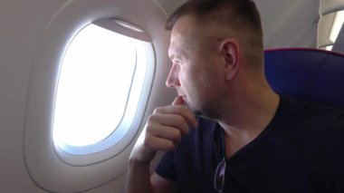 Boynunda güneş gözlüğü olan düşünceli bir erkek yolcu gündüz uçuşu sırasında düşünceli bir şekilde uçak camından bakar, mavi aksanlı uçak kabininde otururken çenesini dinlendirir.
