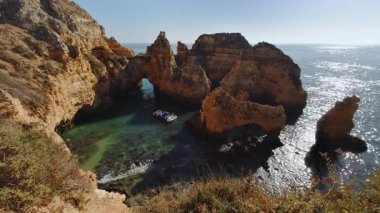 Kristal berraklığında turkuaz sulardan yükselen dramatik altın kireçtaşı kemer ve kaya oluşumlarının yer aldığı nefes kesici kıyı manzarası, ziyaretçilerin muhteşem jeolojik harikalar arasında tekneyle keşif yaptıkları büyüleyici bir doğal koy oluşturuyor.