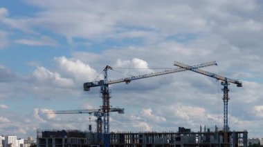 Kule vinçleri, güzel mavi gökyüzünün altında bir apartman inşa eder. Müstakbel ev sahipleri için emlak geliştirme sürecini gösteren beyaz tüylü bulutlarla birlikte.