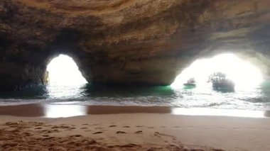 Portekiz 'in Algarve bölgesindeki ünlü Benagil Deniz Mağarası' nın göz kamaştırıcı iç manzarasında doğal tavan penceresi, altın kum plajı ve turkuaz suları, rehberli gezinti sırasında deniz kemerlerinden giren turist botları yer alıyor.