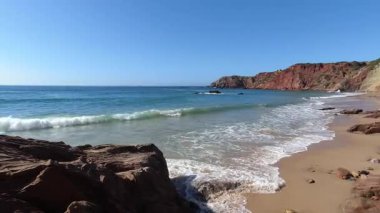 Gök mavisi okyanus dalgalarının büyüleyici manzarası Portekiz 'in batısındaki Algarve kıyıları boyunca uzanan belirgin kızıl kayalıklarla çevrili altın kumlu plajlara süzülüyor. Açık mavi gökyüzü altında yaz tatili için mükemmel.