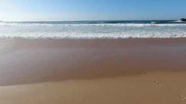 Nazik turkuaz okyanus dalgalarının açık mavi gökyüzünün altında altın kuma vurduğu güzel Portekiz plajı yaz tatili için mükemmel sakin kıyı manzarası yaratıyor.