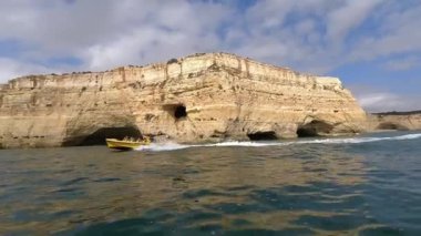 Stunning footage of yellow speedboat navigating along dramatic golden limestone cliffs of Algarve Portugal approaching famous Benagil sea caves and grottos during popular coastal tour showcasing natural rock formations shaped by Atlantic waves