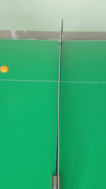 Turuncu masa tenisi topu açık yeşil masa tenisi yüzeyinde kapalı spor salonunda dinleniyor. Eğlence oyunları için profesyonel spor ekipmanları