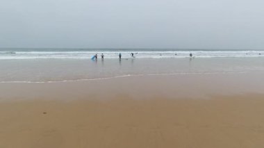 Portekiz 'deki altın kumlu sahilin atmosferik açılı manzarası. Uzak sörfçüler sisli okyanus sularına göğüs gererek sisli ufka karşı siluetler yaratıyor. Hafif dalgalar geniş kıyı şeridine doğru yuvarlanıyor.