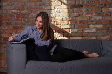 Mavi tişörtlü gülümseyen kadın kanepede uzanıyor ve kalemle deftere yazıyor.