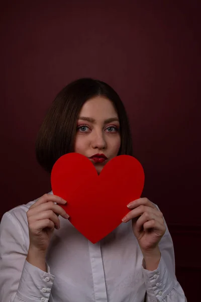 Short-haired woman on red background holding red heart shape in front of her chin