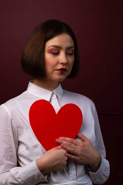 Dreamy short-haired woman on red background holding red heart shape in front of her chest with closed eyes