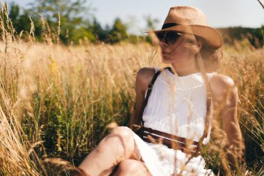Özgürlüğün ruhu. Bluzlu, şapkalı ve güneş gözlüklü çekici bir kız mavi gökyüzünün arka planında sahada duruyor. Yaz tatili, seyahat. Bohem, modern hippi tarzı.