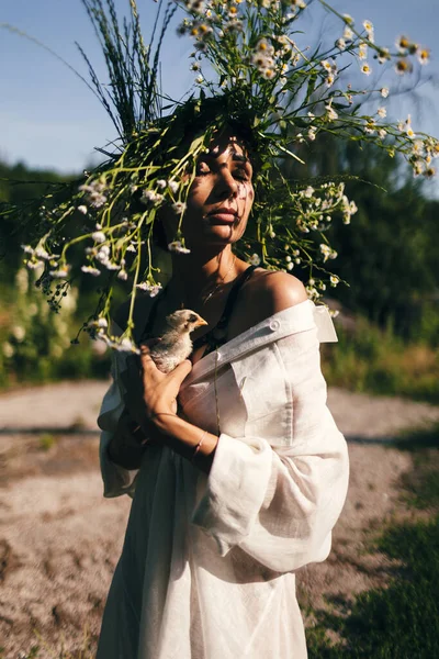一位身穿白色太阳衣 头戴雏菊花环的年轻女子 在森林的背景下 在夕阳的映照下 手中握着微微的颤音 自由概念 — 图库照片