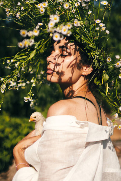 A beautiful young shuman in a wreath of daisies on her head and holds chick in her hands.