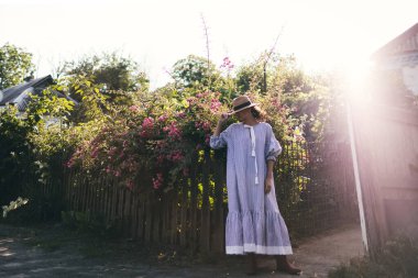 Romantik giyinmiş genç yetişkin güzel bir kadın kırsal bir parkta dinleniyor ve başka tarafa bakıyor. Beyaz şeffaf elbisesi ve hasır şapkasıyla bahçede yürüyen sarışın Paris tarzı bir model..