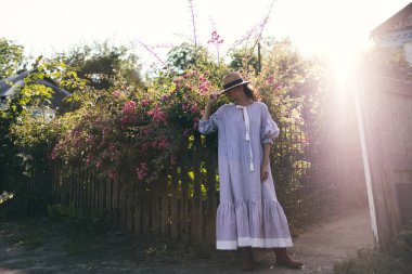 Romantik giyinmiş genç yetişkin güzel bir kadın kırsal bir parkta dinleniyor ve başka tarafa bakıyor. Beyaz şeffaf elbisesi ve hasır şapkasıyla bahçede yürüyen sarışın Paris tarzı bir model..