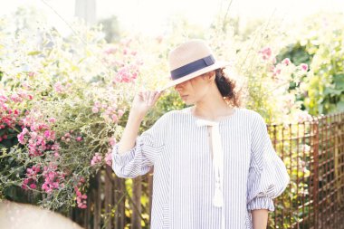 Romantik giyinmiş genç yetişkin güzel bir kadın kırsal bir parkta dinleniyor ve başka tarafa bakıyor. Beyaz şeffaf elbisesi ve hasır şapkasıyla bahçede yürüyen sarışın Paris tarzı bir model..