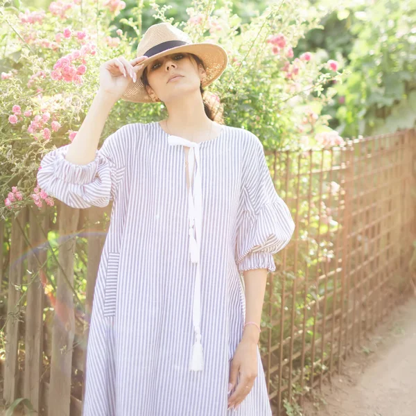 在一个乡村公园里 穿着时髦的年轻美丽的成年女子在一边看一边放松 布隆德 巴黎风格的模型 身穿白色透明的连衣裙 头戴草帽 走在花园旁边 — 图库照片
